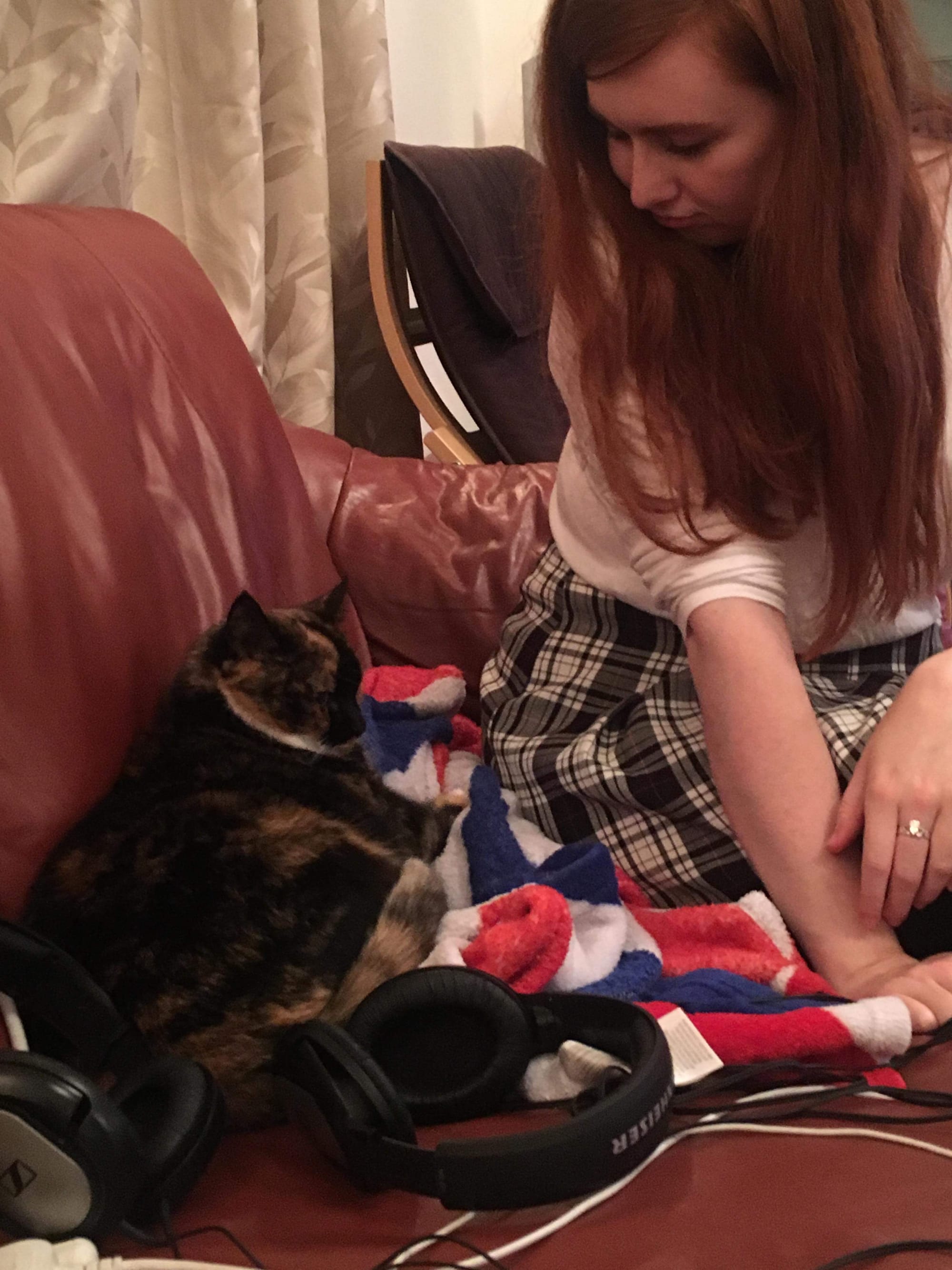 A person with long red hair sits on the edge of a sofa seat, looking behind them at a black and ginger cat who is curled up for a nap