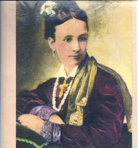 A colourised image of a woman in a velvet dress and large crucifix necklace. Her hair is tied up with flowers in it, and she is looking away from the camera.