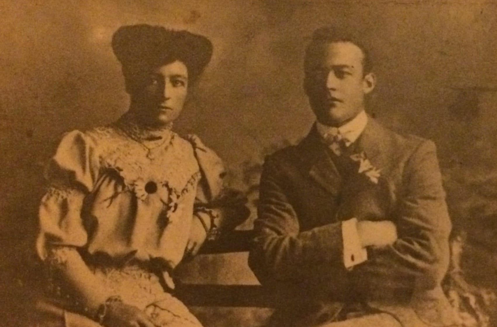 A woman and man look sombrely at the camera, sat on a bench, in formalwear. 