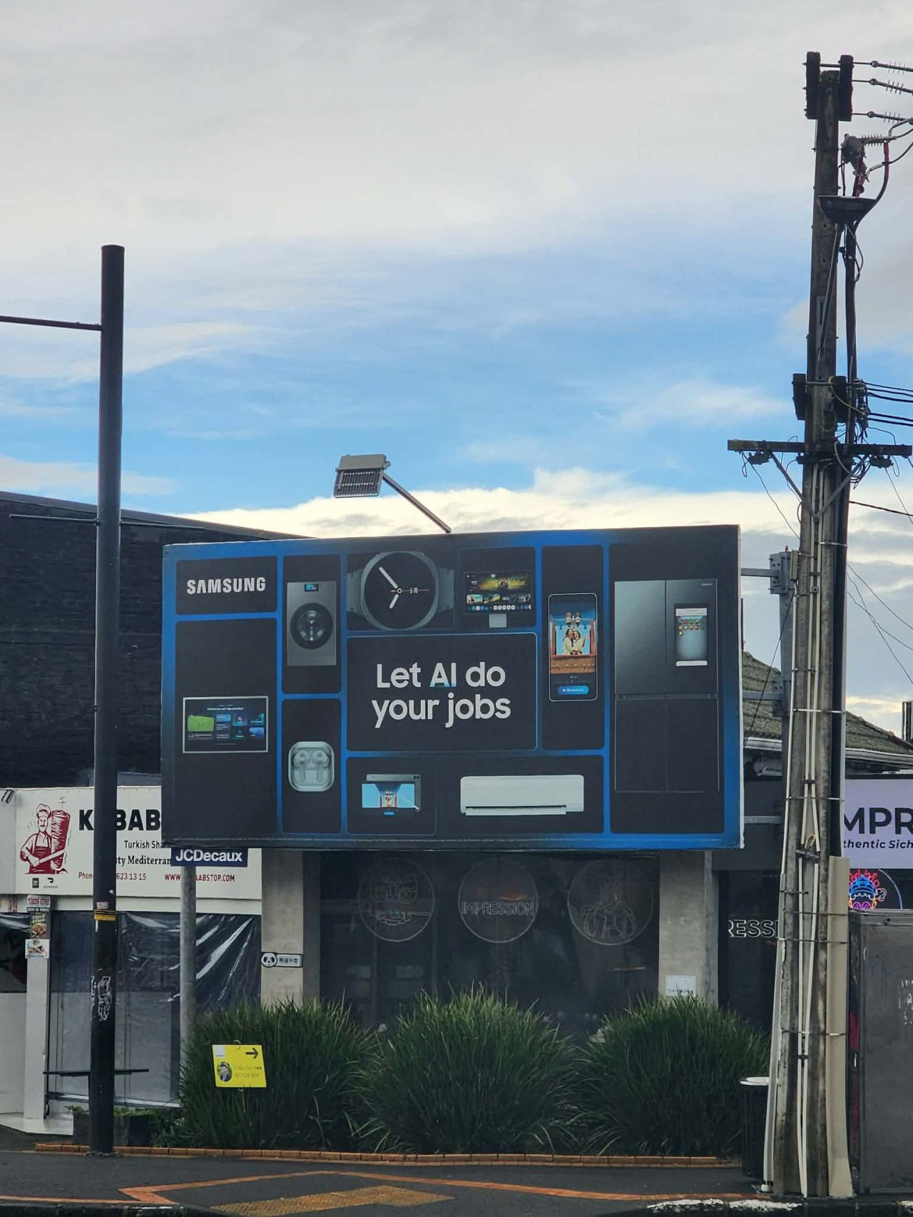 A billboard sits above a kebab restaurant. It reads 'Let AI do your jobs' and features photos of tech devices such as earphones and a computer.