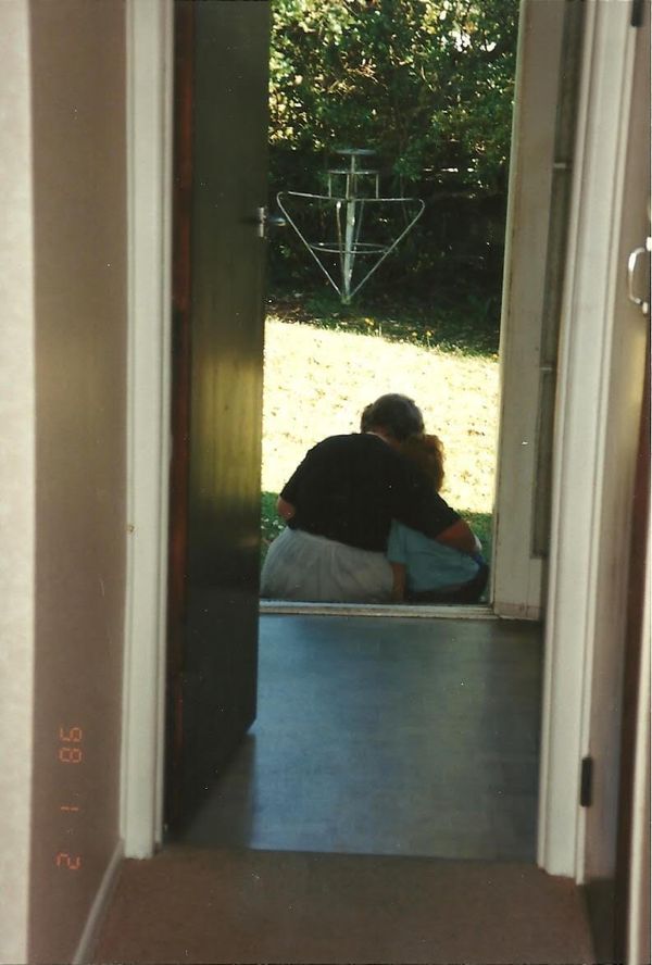 A photo down a hallway of an old woman and her 3-year old granddaughter cuddling while sat on the doorstep, looking at the sunny garden.