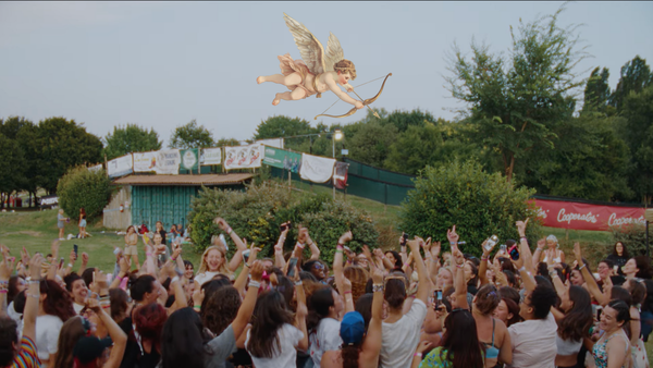 A screengrab of a group of 100 girls, all Harry Styles fans, jumping and singing in a field before a concert. An illustration of Cupid is aiming down at the group.