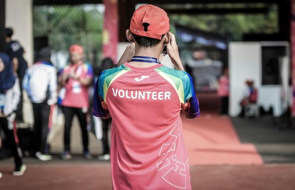 A man in a bright jersey with "VOLUNTEER" on the back faces away, adjusting his cap in a busy sports event setting. Others in similar attire are in the background.