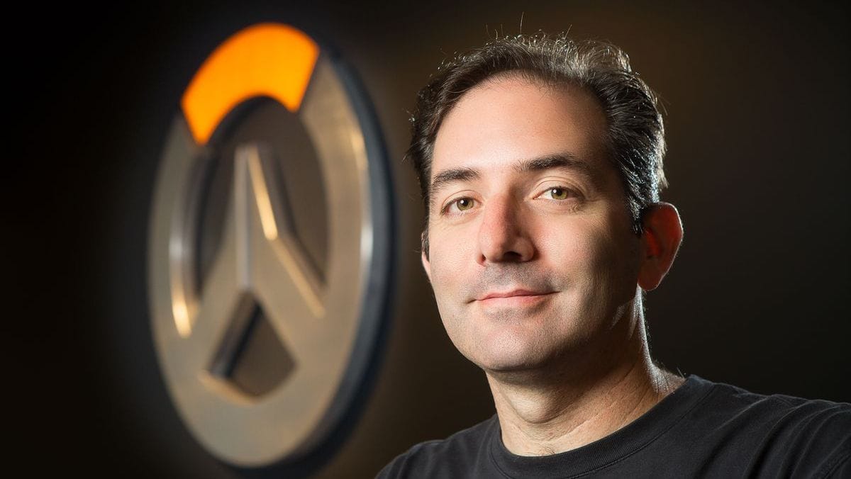 Jeff Kaplan looking at the camera with the Overwatch logo on a wall out of focus behind him.