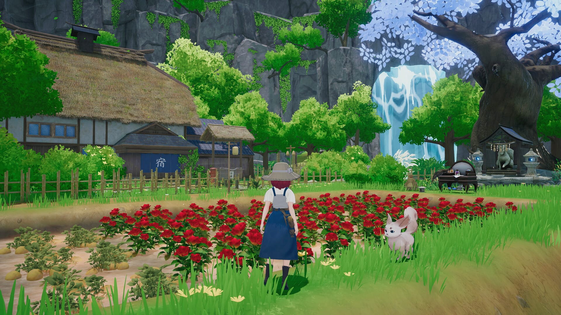 A woman stands next to a rosebed in front of a thatched cottage, waterfall and trees in the distance.