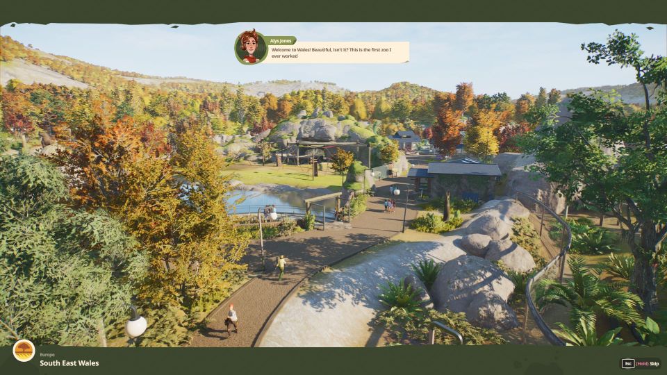 The camera floating above a tree-covered, landscaped zoo. "Welcome to Wales!" says the character Alys Jones. "Beautiful, isn't it?"