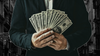 A composite image of a faceless person in a suit, holding a fan of $100 bills, overlaying a photograph of New York City.