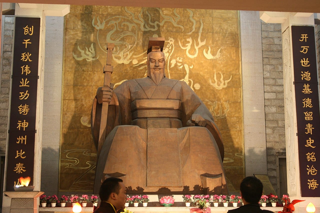 Estatua de madera del Emperador Amarillo, Huangdi, sentado en un trono dentro de un recinto moderno. Está vestido con túnicas tradicionales y sostiene un cetro, frente a un fondo dorado con motivos de nubes estilizadas. A la izquierda, una columna con inscripciones chinas acompaña la escena, mientras que en primer plano, se observa a una persona pasando, proporcionando una escala a la imponente estatua. La atmósfera es de reverencia y la estatua actúa como un punto focal para la contemplación y el respeto por el histórico Emperador Amarillo.