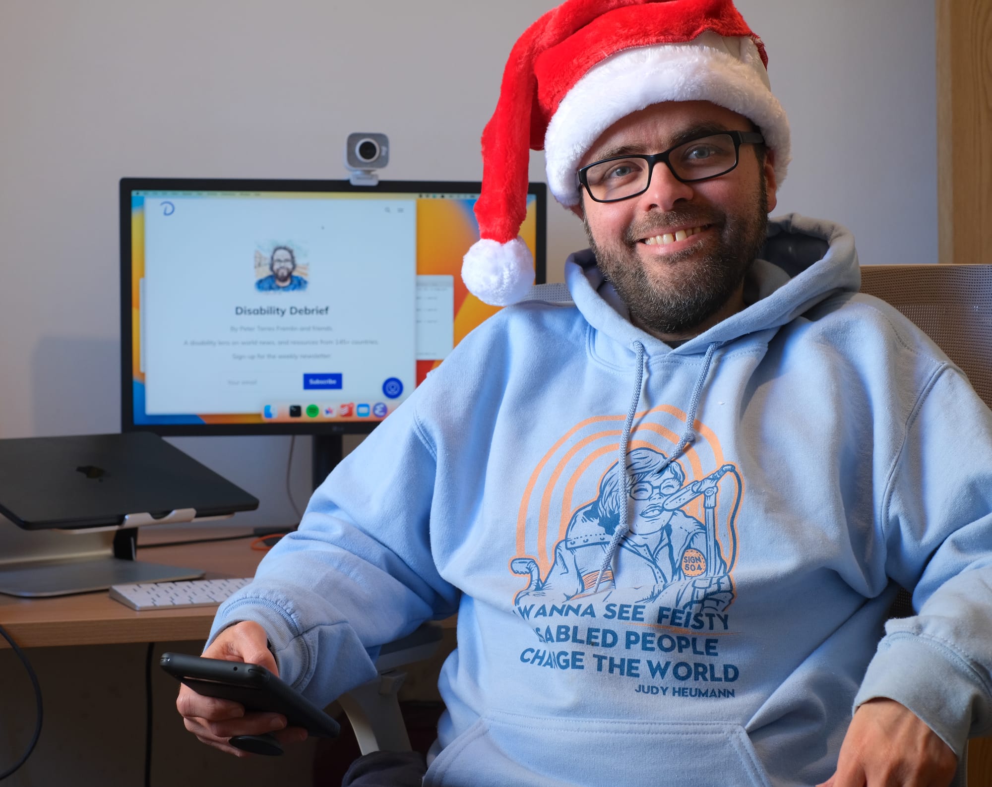 Picture of Peter at his desk, smiling and wearing a Santa hat. Peter is white, has glasses and a beard with speckles of grey. He's wearing a Judy Heumann hoodie that says 'I wanna see feisty disabled people change the world.'