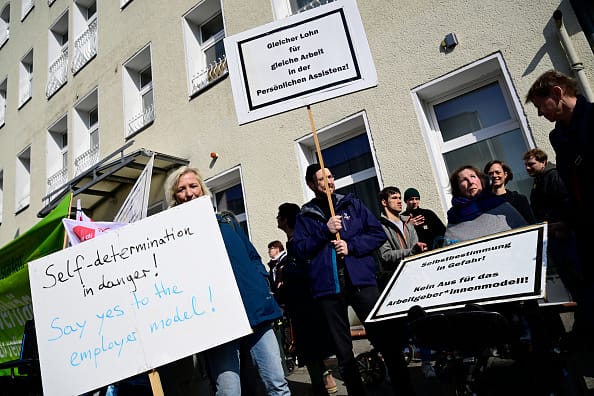 Photograph of a group of demonstrators holding placards during a protest. Some of the demonstrators are wheelchair-users; several of them hold placards which read "Self-determination in danger! Say yes to the employer model!", "Selbstbestimmung in Gefahr! Kein Aus für das Arbeitgeber*innenmodell!" and "Gleicher Lohn für gleiche Arbeit in der Persönlichen Assistenz!".