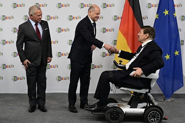 Photograph of Chancellor Olaf Scholz shaking hands with Nawaf Kabbara, while King Abdallah II looks on smilingly. The men are dressed smartly in dark suits and Nawaf Kabbara is a wheelchair-user. In the background behind them, there is a German flag and EU flag in front of Global Disability Summit branding.