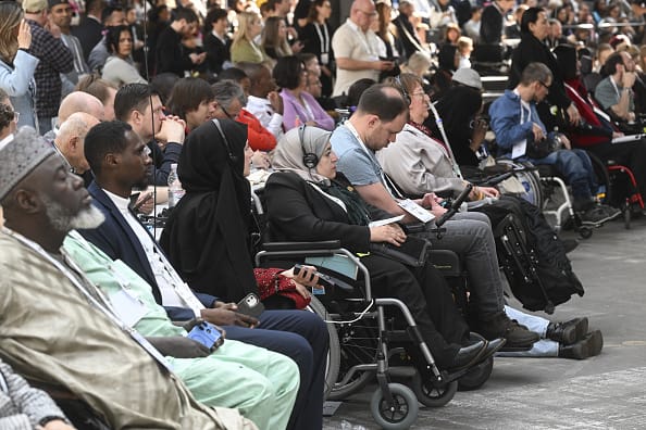 Photograph of a big crowd of delegates sit in the audience. In the front row, there are three Black men, and beyond them sit two women wearing headphones over their headscarves. Many of the delegates are wheelchair-users and one is using a ventilator. The delegates all wear a look of concentration as they gaze into the same direction.