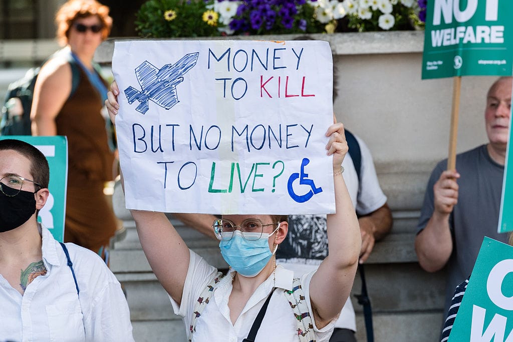 A photo of a young white person who wears a facemask holding a sign above their head that reads "Money to kill but no money to live". The sign shows both a hand-drawn rocket and a blue wheelchair symbol. In the background, other protestors hold banners and signs of their own.