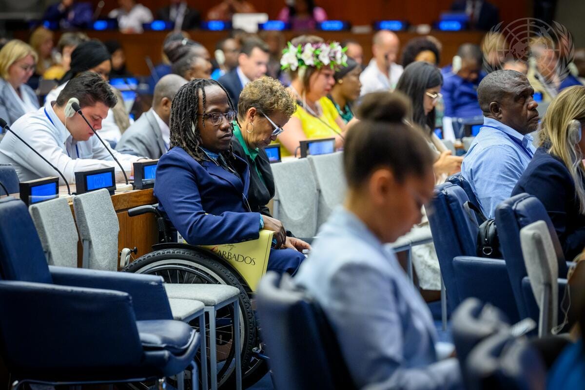 A crowded view of around thirty delegates sitting in rows in a large meeting room. In focus in the foreground, a Black man with locs is looking directly at the camera with a look of scepticism. He wears glasses, a dark blue suit and sits in a wheelchair, and his arm is resting on a file which reads "Barbados". Around him are delegates of different ages, genders and ethnicities, some of them wearing earpieces and all of them wearing a look of concentration. One woman, in a yellow top, wears a flower crown on her head. There are also a number of empty seats.