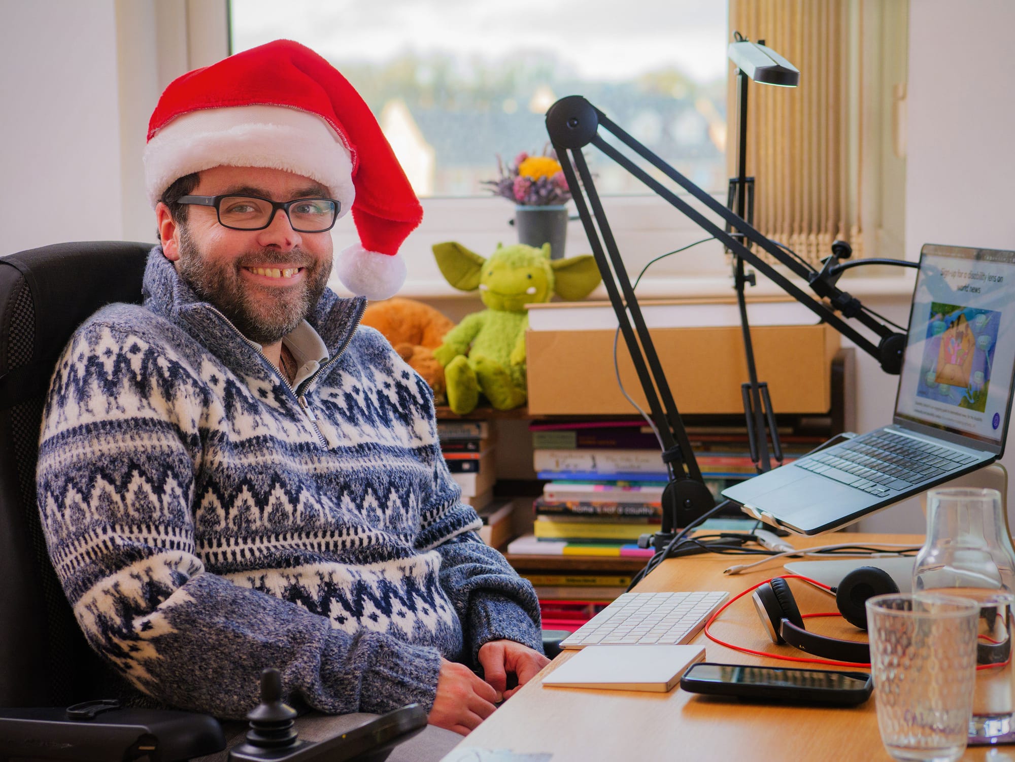 A photo of Peter, a white man with a dark beard, blue eyes and dark-framed glasses, wearing a Santa hat and smiling at the camera. He is wearing a Fairisle blue-and-white sweater, and is seated in his power wheelchair at his desk in his ergonomic home office. His laptop is open on the Disability Debrief sign-up page, and he has a scar on his nose from a recent injury. In the background of the photo, books are piled high to a window and upon them sit a couple of soft toys.