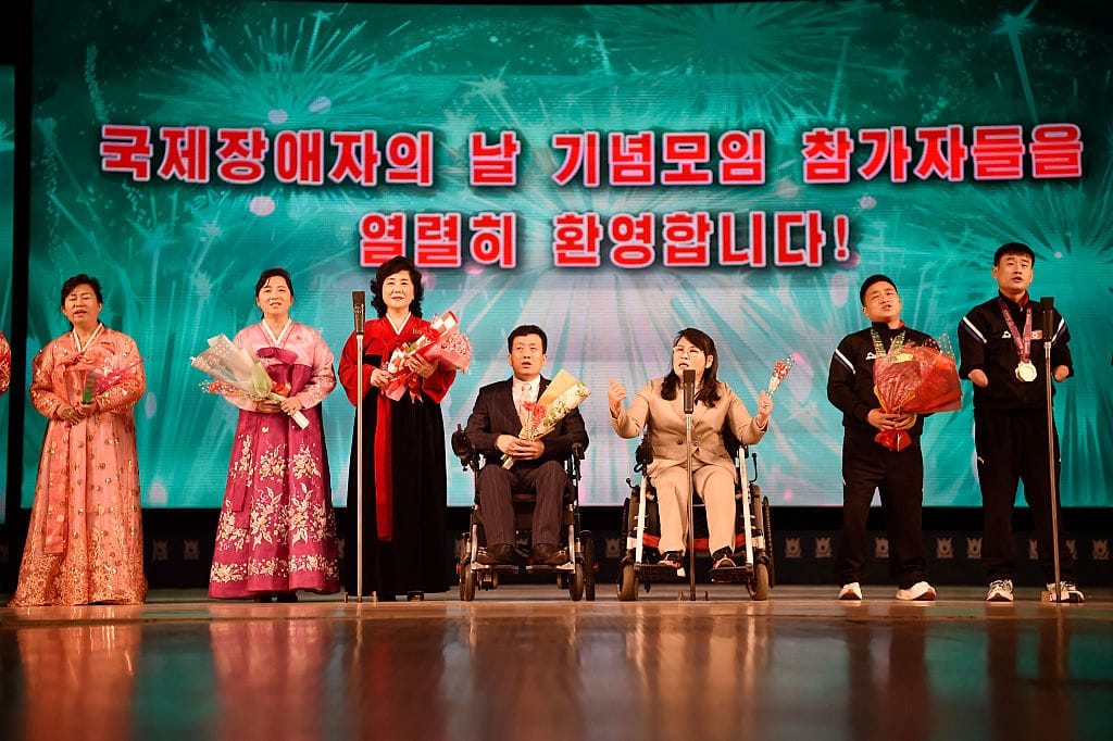 A brightly coloured photograph of seven men and women, several of whom are wheelchair-users or visibly disabled. They are side by side on a kitsch, carefully choreographed stage backed by a screen with Korean text marking the International Day of Persons with Disabilities. Three women wear colourful traditional dresses, a man and women in the centre use wheelchairs and wear suits, two young men in sports clothing wear medals around their necks. Each person is holding a bouquet of flowers, and most of them have their mouths open or eyes closed, as if singing into their microphones.