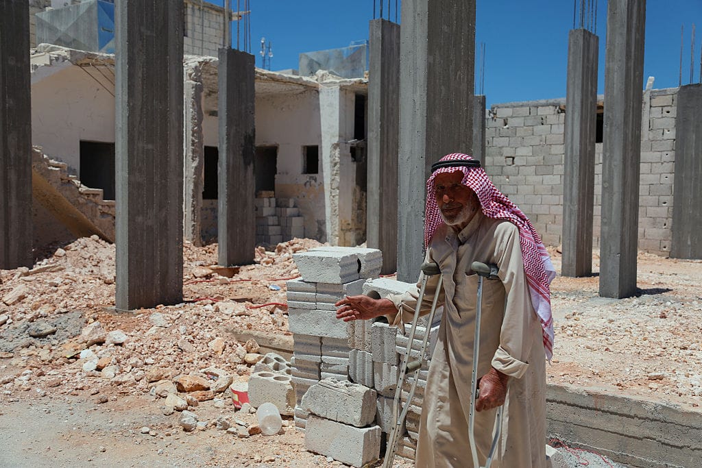 A photo of a late middle-aged man supporting himself on crutches and wearing a red keffiyeh as he stands in front of a construction site. The man wears a slight smile, and one of his hands appears to be gesturing behind him. Behind him, nine brand new grey concrete supportive columns stand tall, awaiting their next stage of construction, and piles of grey concrete bricks lie in the sandy-coloured rubble. The original building has been so badly damaged that only a couple of its external walls and one internal staircase remain. The photo has been taken in bright sunshine, and in the background the sky is a bright blue.