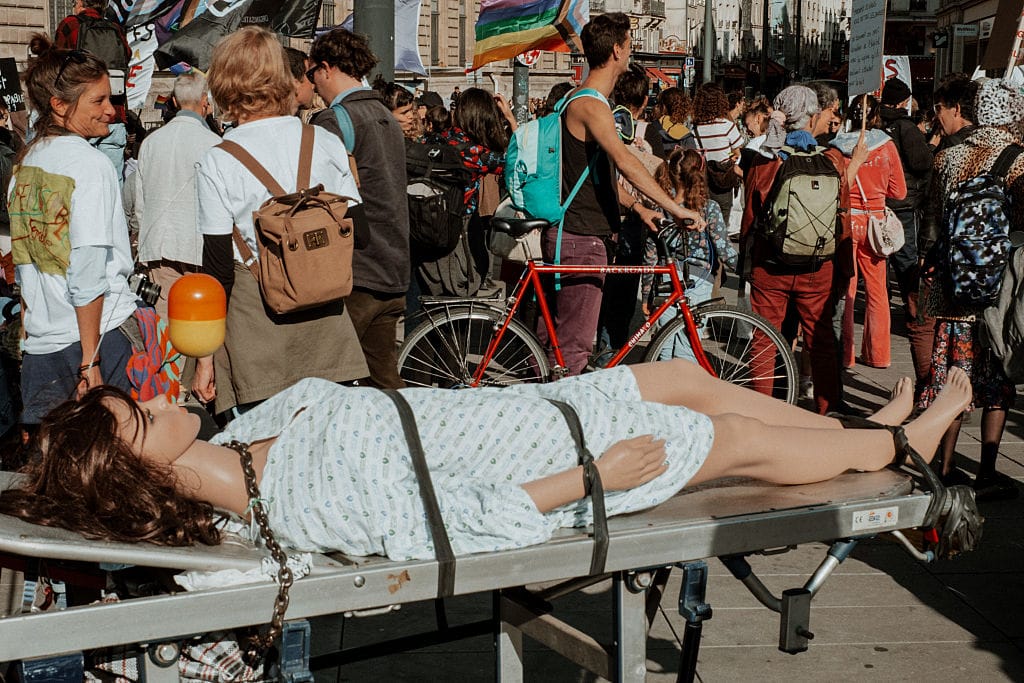 A photo of a female mannequin who is strapped to a stretcher and wearing a hospital gown, at a protest. She is strapped to the stretcher by a metal chain around her neck and black fabric ties around her stomach, hands and feet. She looks young and has long brown hair, and her facial expression is vacant and pointed towards the sky. A giant orange-yellow pill hangs suspended above her head. In the background, a large crowd of protesters of different ages have their backs to us. Some hold placards, and an LGBTQ+ Pride flag is flying in the air.
