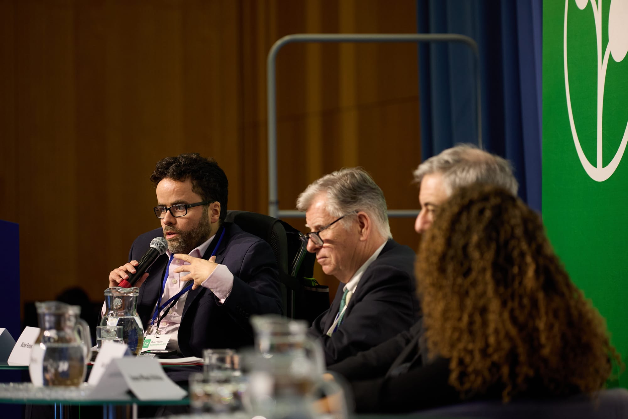 A photo of Peter, a white man with dark hair, beard and glasses, wearing a pink shirt and navy blue suit and talking into a microphone as he sits beside three other people on a panel. Peter is in focus, while two other white men and a woman with curly hair are slightly out of focus, as are the jugs of water, glasses and name badges which sit on the table in front of them. In the background, there is a wood panelled wall, a blue curtain, and a corner of a large green Zero Project logo.
