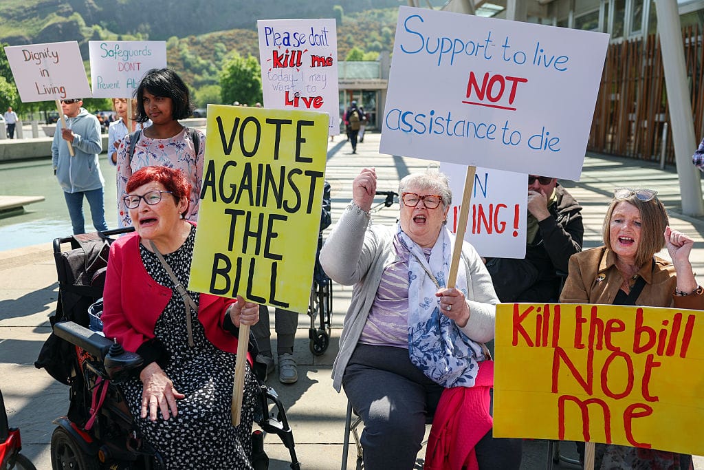 A photo of a group of demonstrators, most of them wheelchair-users, protesting passionately while holding handmade placards which oppose an assisted dying bill. In the foreground, three older white women wheelchair-users have their mouths open in outcry and their fists raised as they hold placards which read "VOTE AGAINST THE BILL", "Support to live NOT assistance to die" and "Kill the bill NOT me." They are in bright sunshine, and behind them a modern square opens onto the Scottish Parliament and a lush green hillside.