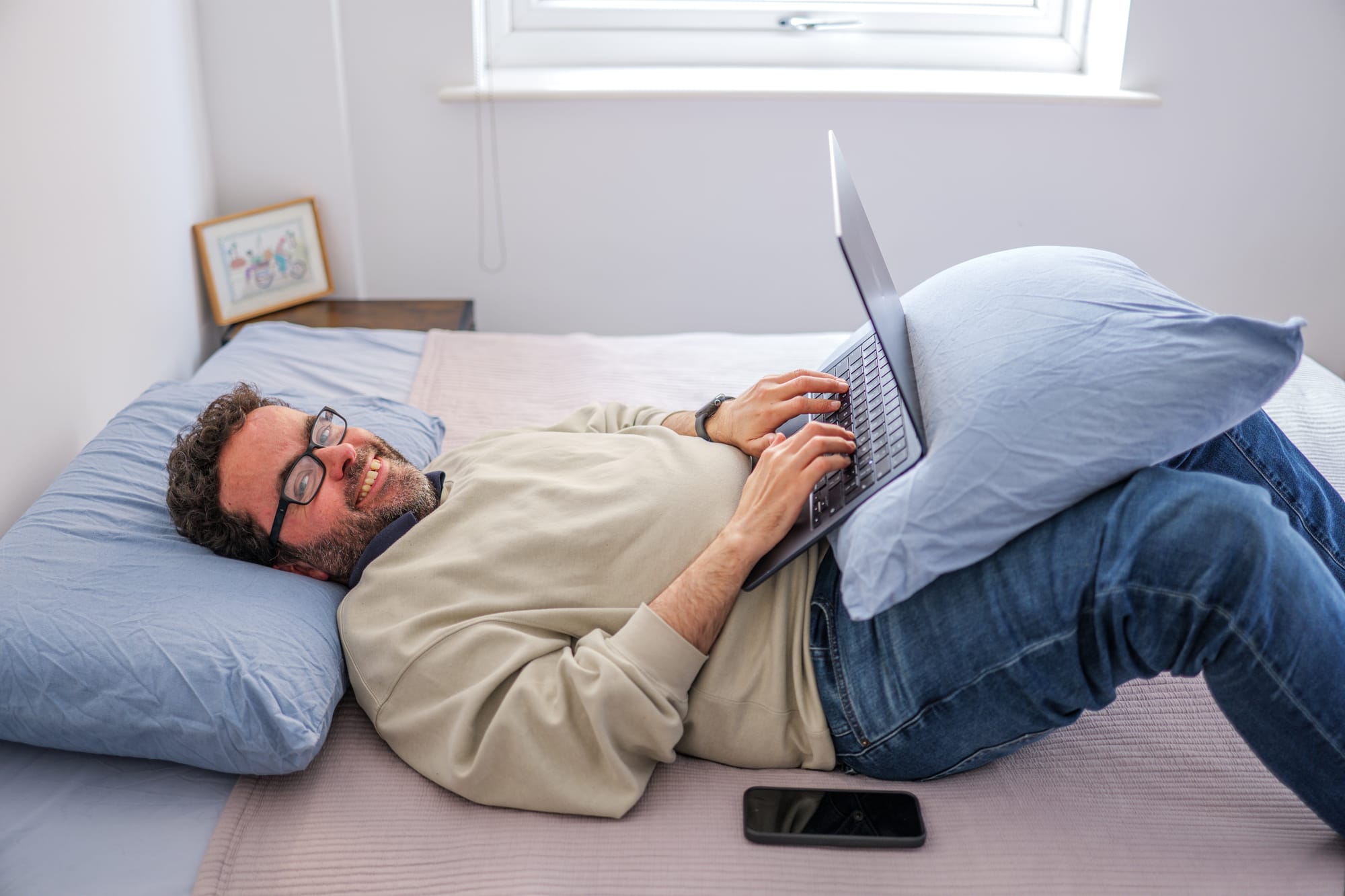 A photo of Peter, lying on his back in bed and using his laptop, which is propped on a pillow against his folded knees. Peter is a white man with dark hair, beard, and glasses, smiling at the camera while his phone rests on the bed beside him. He is wearing a grey-green jumper, dark blue jeans, and his neat bedsheets and pillows are blue and beige. In the background of the photo, a framed illustration sits on a dark wood bedside table, and to its right, we can see the bottom of a bright white window frame.