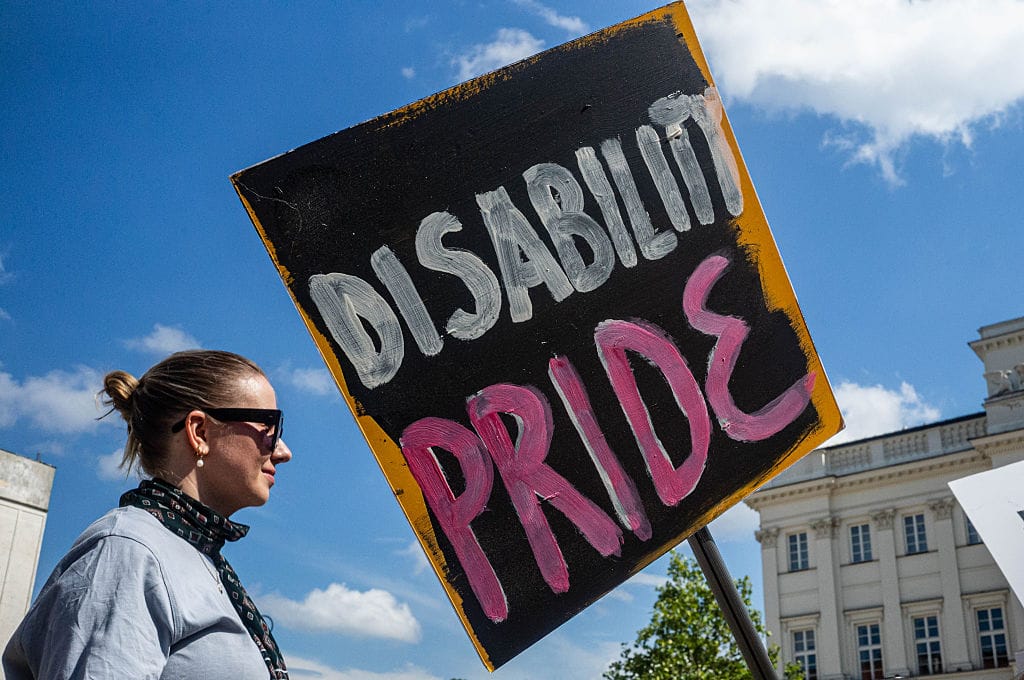 A blonde woman faces a placard reading DISABILITY PRIDE. The sign is held on a stick at a slightly crooked angle and PRIDE is in pink. The woman, visible in profile, has sunglasses and a gentle smile. Behind them are a blue sky with some clouds and an impressive building in neoclassical-style.