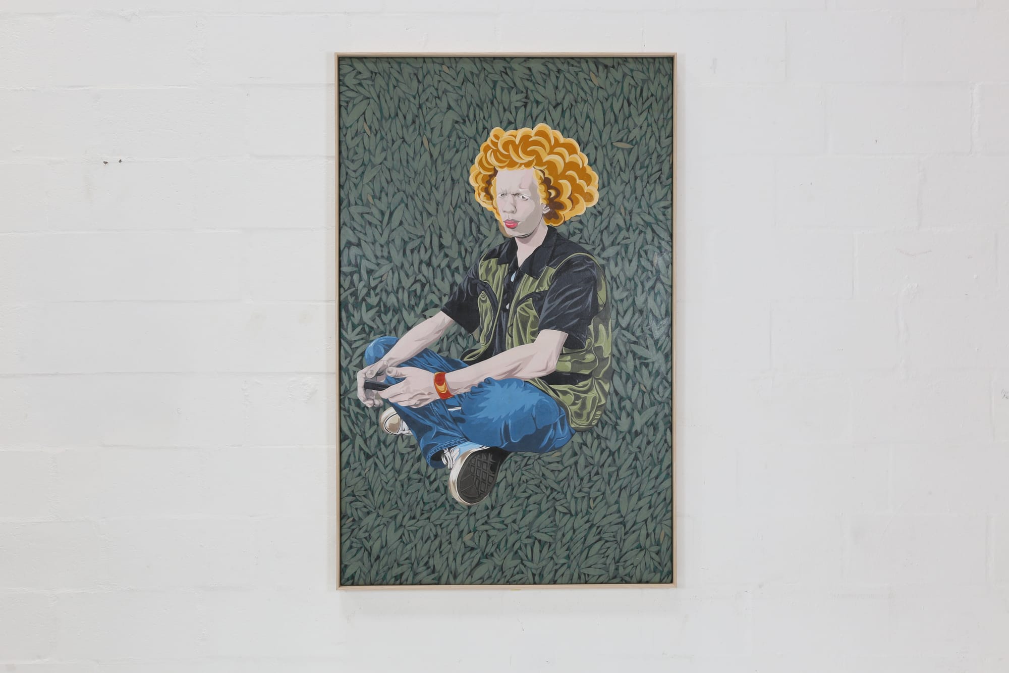 Picture of a painting hanging on a white wall. In the painting, ayoung man with albinism sits cross-legged against a densely patterned backdrop of layered leaves, fading from deep emerald, green to lighter shades of green. He wears a black shirt adorned with intricate gold detailing, paired with blue trousers and black-and-cream Converse sneakers. Voluminous burnt-orange curly hair frames his pale skin, creating a striking contrast with the lush green background. His relaxed pose and direct presence evoke a sense of confidence, individuality, and quiet strength.