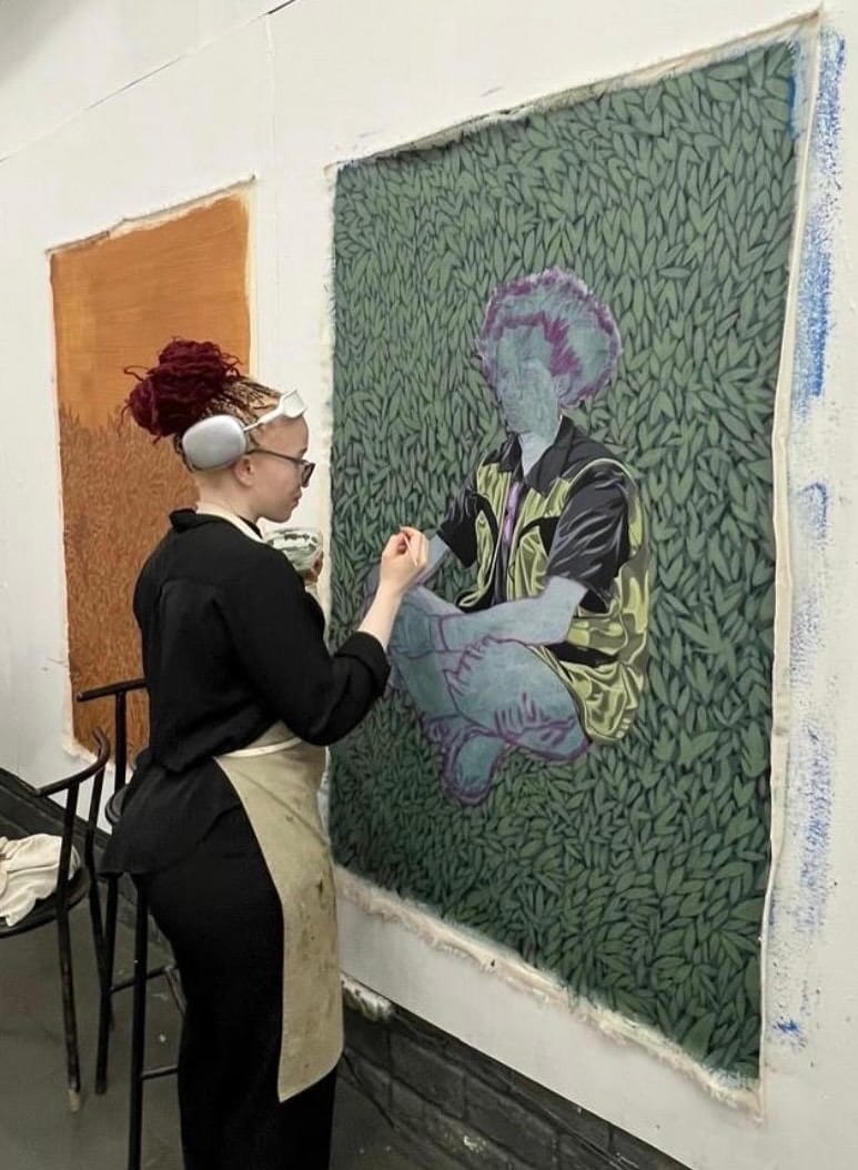 Photograph of a young woman with albinism, dressed in a black shirt, black pants, and a beige apron, stands beside a large artwork, appearing to work on the piece. The painting depicts a cross-legged seated figure against a patterned green backdrop. The face and legs are blank, but a large outline of hair is visible.