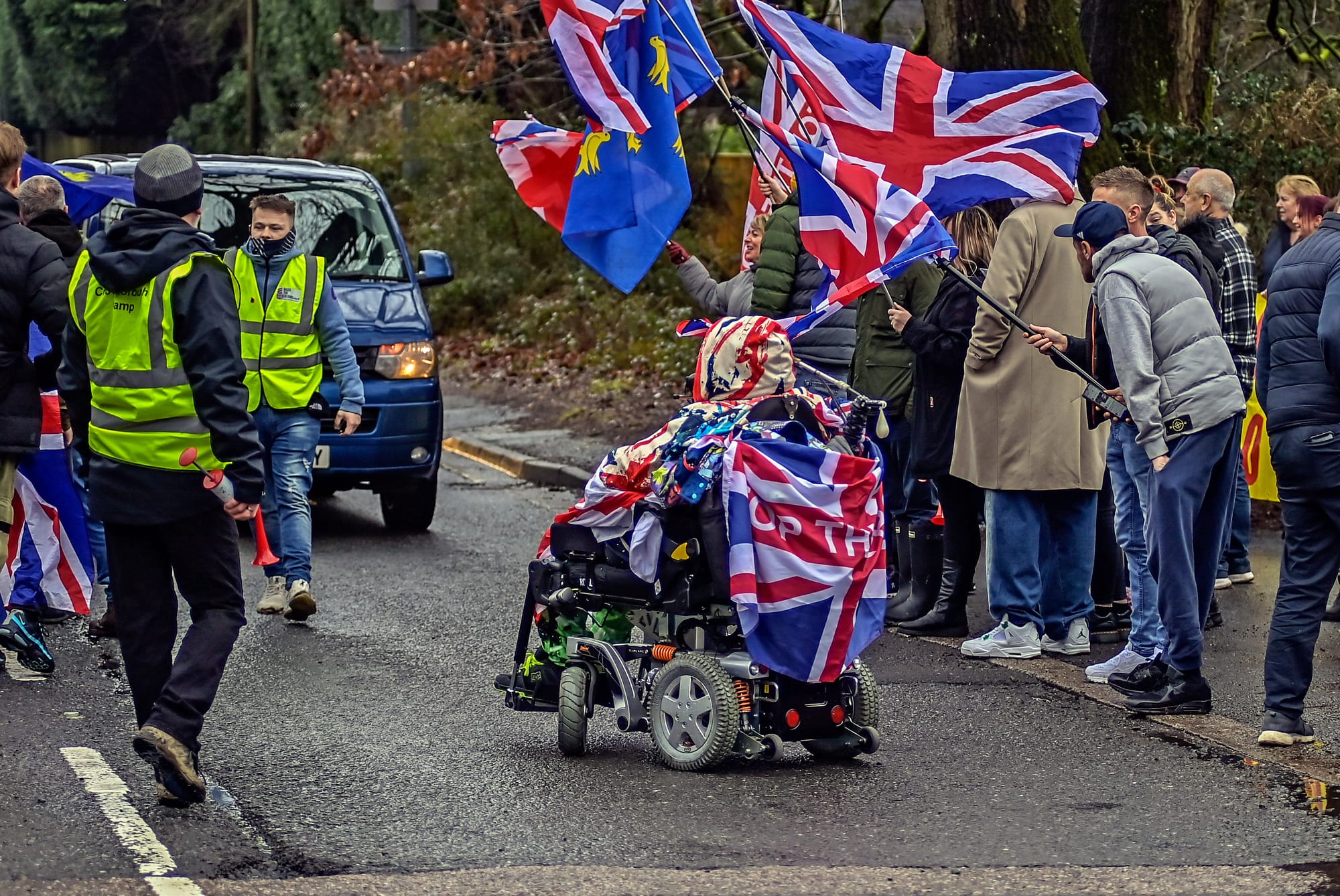 A photo of a roadside protest, where a powerchair-user draped in a Union Jack flag is blocking an oncoming blue van. Photographed from behind, the powerchair user is in the middle of the road and to his right, on the pavement, a group of more than a dozen people are zealously waving Union Jack flags and also facing the direction of the oncoming van. To their left, two men are wearing yellow reflective vests.