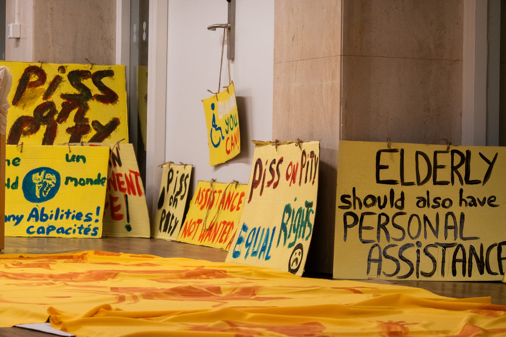 A photo of eight huge handmade signs which have been painted with bold lettering onto yellow cardboard and propped against the exterior walls and closed door of a conference room. Three of the signs read "PISS ON PITY" with big black letters, and another prominent one in the foreground reads "ELDERLY should also have PERSONAL ASSISTANCE." Some of the smaller signs read "ASSISTANCE NOT WANTED" and "EQUAL RIGHTS," one has a hand-drawn wheelchair symbol, and another has a simple sad face.