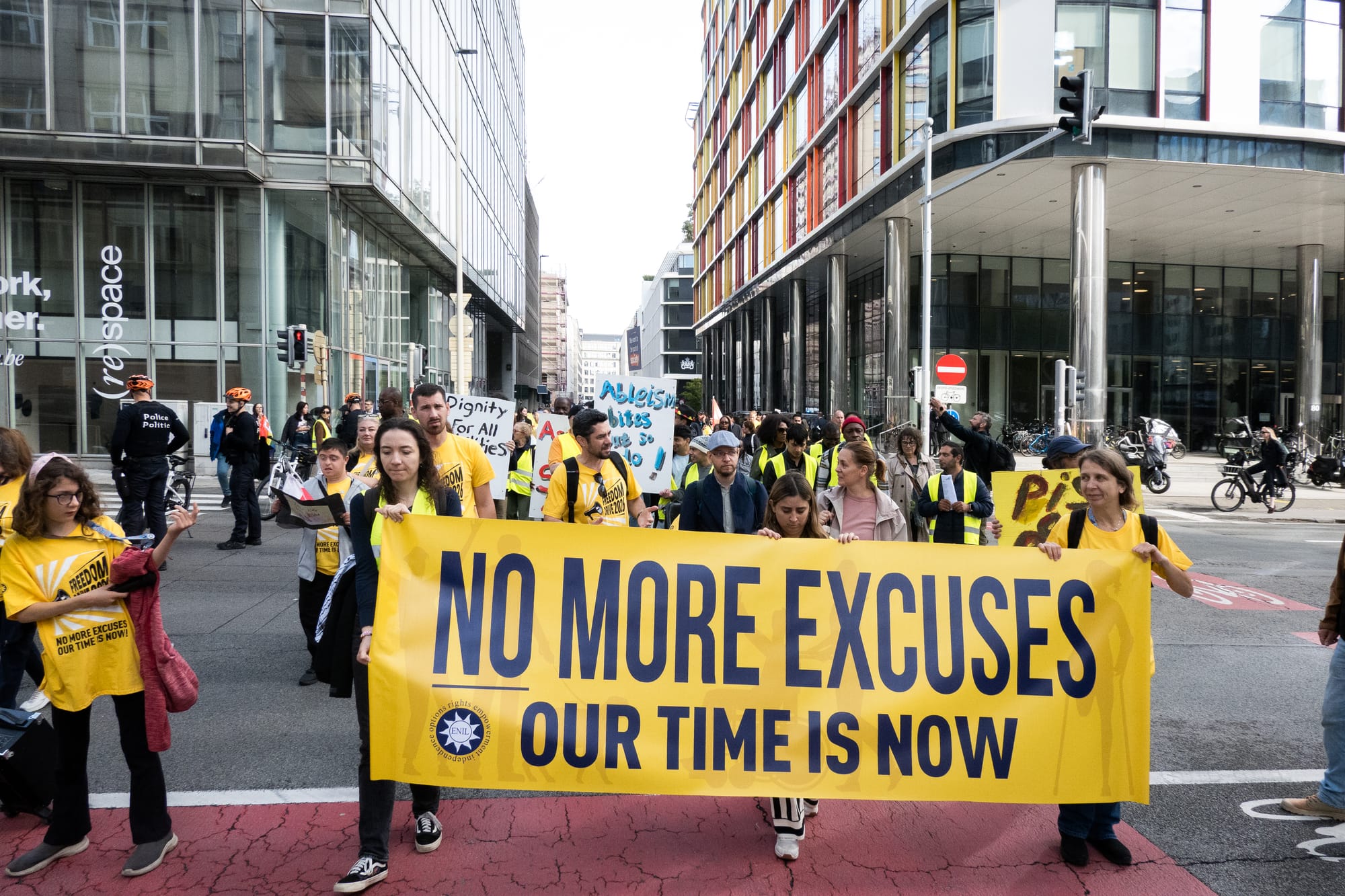 A photo of a large crowd marching across a junction in a modern city centre, holding a huge horizontal yellow banner which reads "NO MORE EXCUSES/ OUR TIME IS NOW". The crowd are a mix of ages and genders, many of them wear yellow t-shirts which read "Freedom Drive 2026" or hi-viz jackets, and a few hold hand-painted placards. In the second row behind the banner are two men talking, one with sunglasses on his yellow shirt and another with a dark jacket and cap. And on the edge of the march are two policemen wearing cycling helmets and stopping traffic.