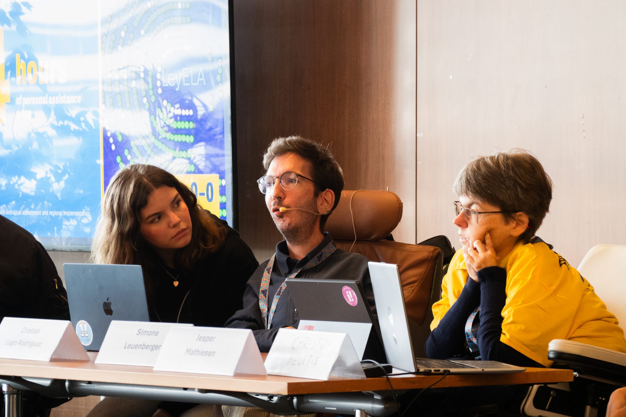 A photo of a young wheelchair-using man speaking into an earset microphone while two women sitting beside him at the conference table lean in and listen intently. The man has short dark hair, wears glasses and has a small frame. They sit on a panel in front of laptops and name badges. The woman on the left of the photo is young, has long hair and wears black clothing, and the woman on the right is middle-aged, a wheelchair-user, has short hair and wears a bright yellow t-shirt.