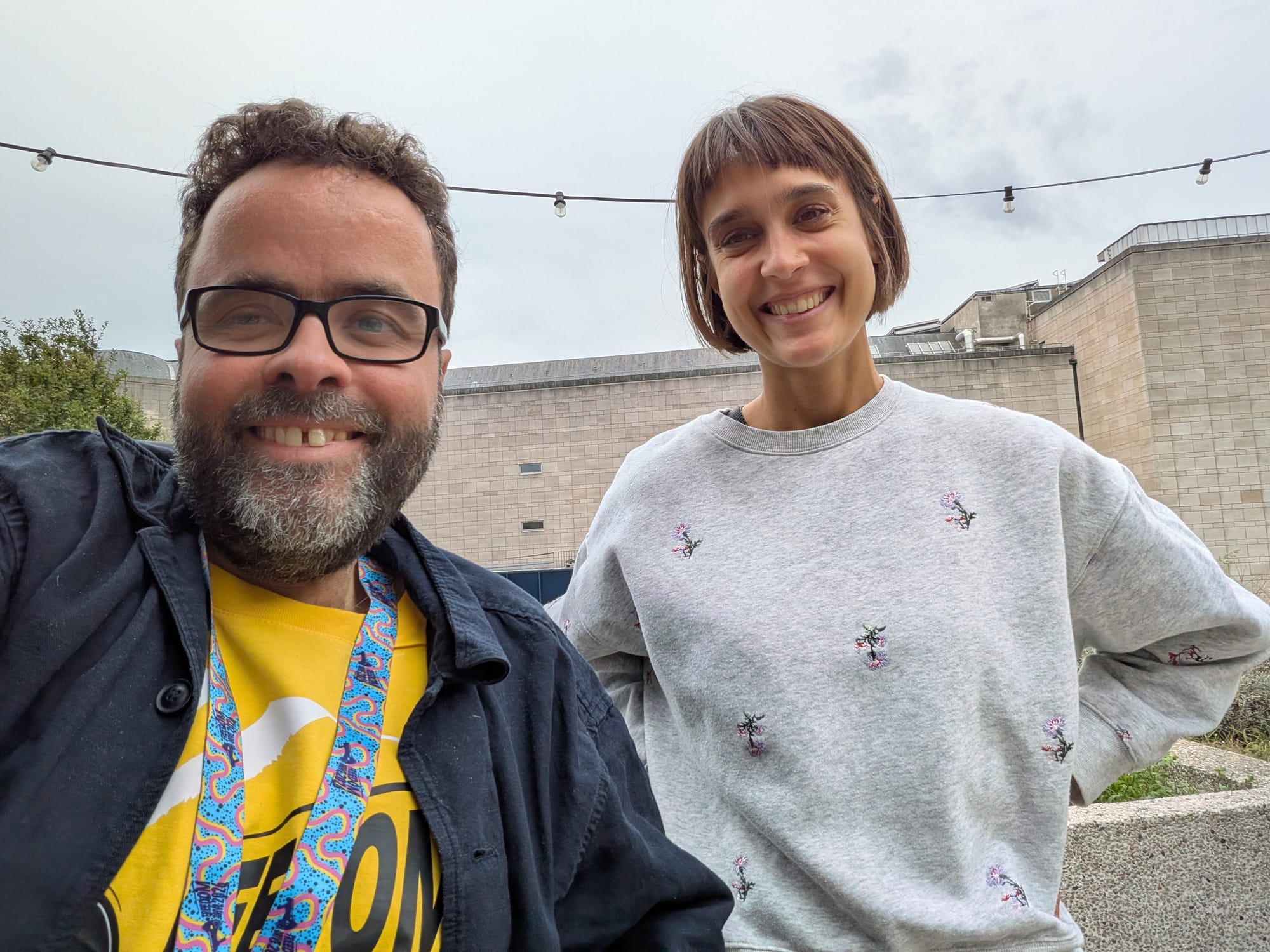 A selfie of Peter and Lea, on a rooftop, both smiling widely. Peter is a middle-aged white man with dark hair, beard, and glasses, and he is wearing a yellow Freedom Drive t-shirt with a dark blue over-shirt and colourful lanyard. Lea is a young woman with an olive complexion, short brown hair, and grey pullover with embroidered flowers. In the background are some globe lights, an imposing grey concrete building and an overcast sky.