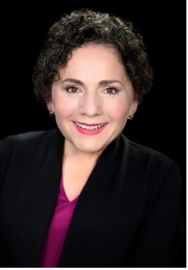 Profile of Yazmine, a white woman with dark hair, looks to the camera, smiling widely. She wears a dark jacket on a dark purple shirt, against a dark background. 