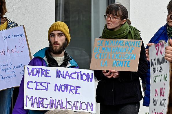 A photograph of two protestors, a man in a wheelchair and a woman standing, holding signs in French. 