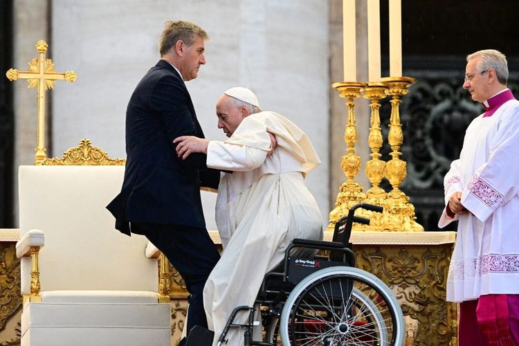 A photograph of Pope Francis being lifted out of his wheelchair by a tall man wearing a dark suit.