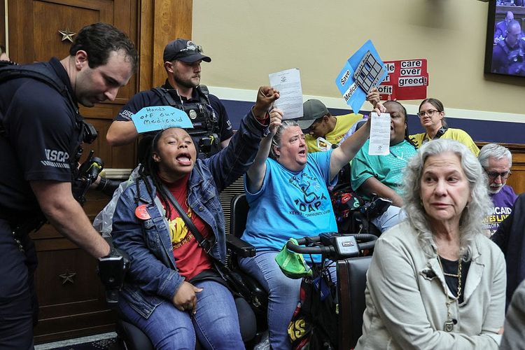 A photo of wheelchair-users protesting in a US government committee room, while two members of the USCP police intervene beside them.