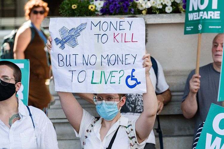 A photo of a young white person who wears a facemask holding a sign above their head that reads "Money to kill but no money to live".