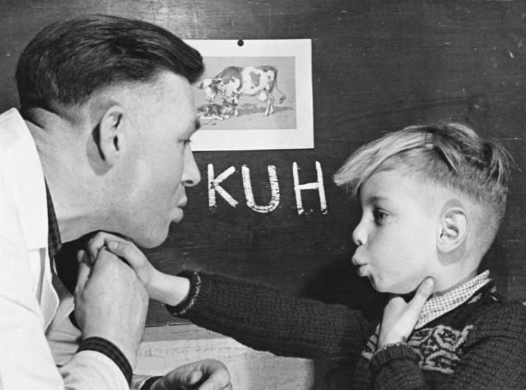 An old black-and-white-photo shows an instructor with a long white coat leaning over a boy, as they both mouth a word.