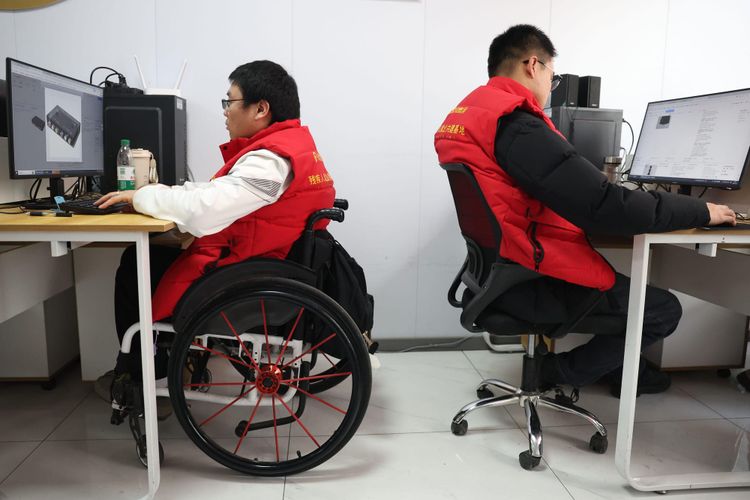 A photo, taken in profile, of two Chinese people, one of whom is a wheelchair-user, sat in front of computers at their two desks in an office.