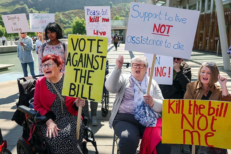 A photo of a group of demonstrators, most of them wheelchair-users, protesting passionately while holding handmade placards which oppose an assisted dying bill.