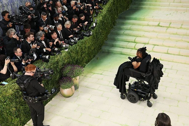 A long-shot photo of a Black powerchair-user posing for the cameras as she is photographed on her own at the bottom of the Met Gala steps.