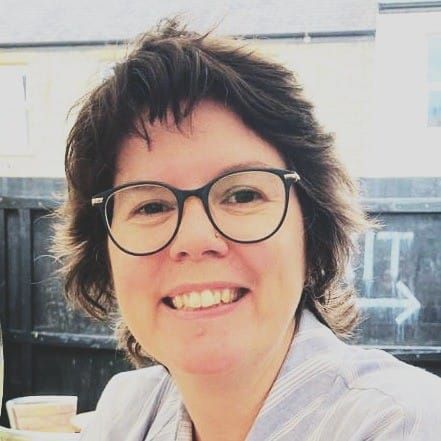 Profile picture of Anna, a white woman with an engaging open smile, glasses with big lenses, and shaggy short hair.