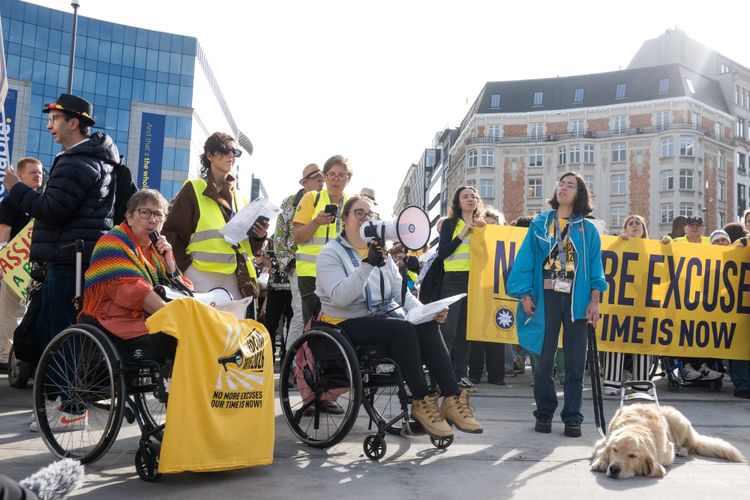 A photo of two wheelchair-using women animatedly speaking into a microphone and megaphone at the front of a protest in a city square.