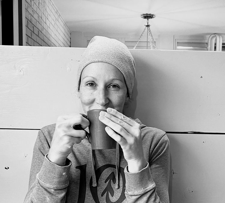 A black and white profile picture of Meritt, a white woman, holding a cup of coffee. She has smiling eyes, a hoodie, and cap covering her hair.