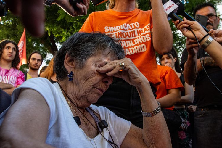 A close-up photo of an older woman seated, covering her eyes with her hand, with a crowd of activists and reporters gathers around her.