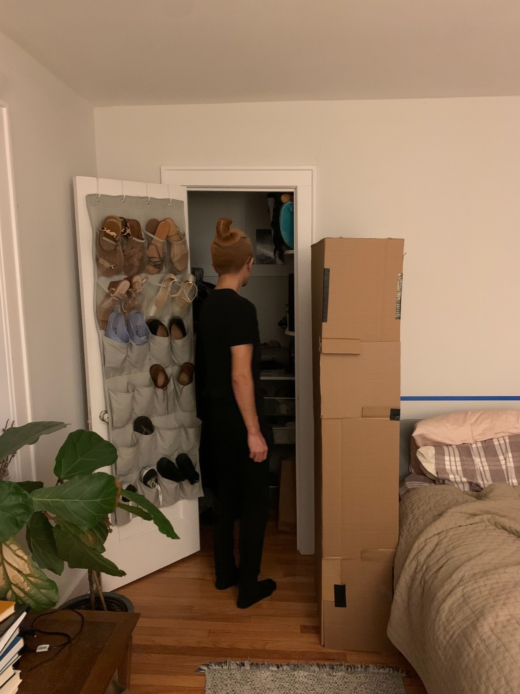 Me standing, back to camera, looking into closet. Open door to the left, pairs of shoes hanging from a over-the-door fabric hanger, cardboard cabinet volume to the right.