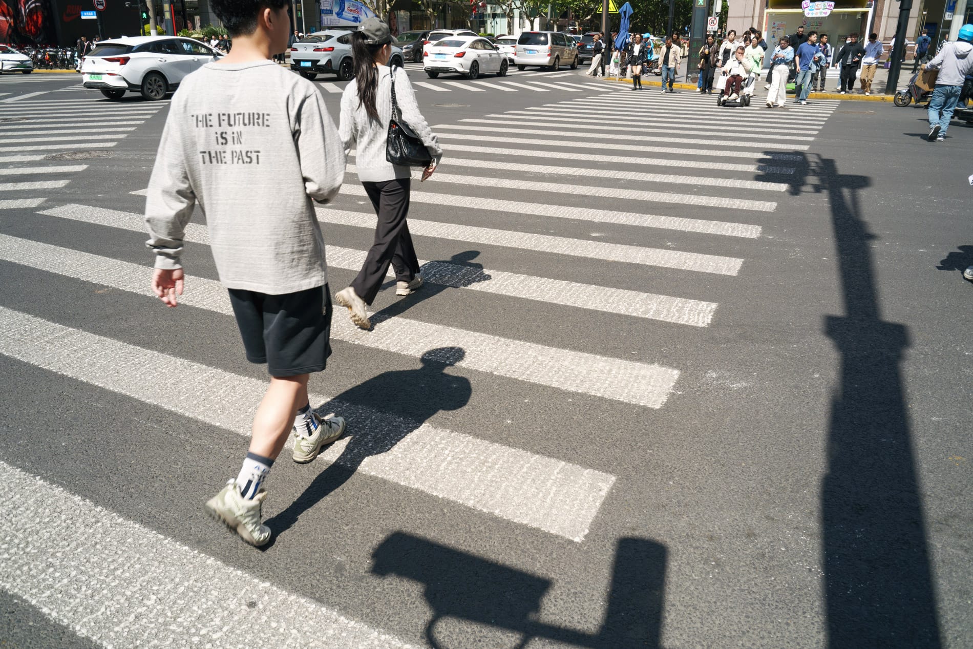 Crosswalk with shadows of surveillance equipment. Walking person with shirt that says on the back, THE FUTURE IS IN THE PAST.