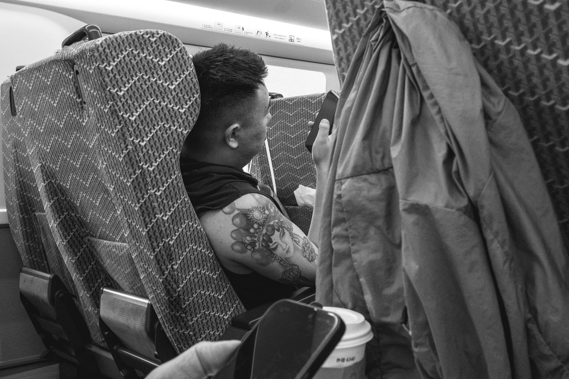 On high speed rail in Hainan. Man in black tank top with large Guanyin tattoo on arm looking at phone.