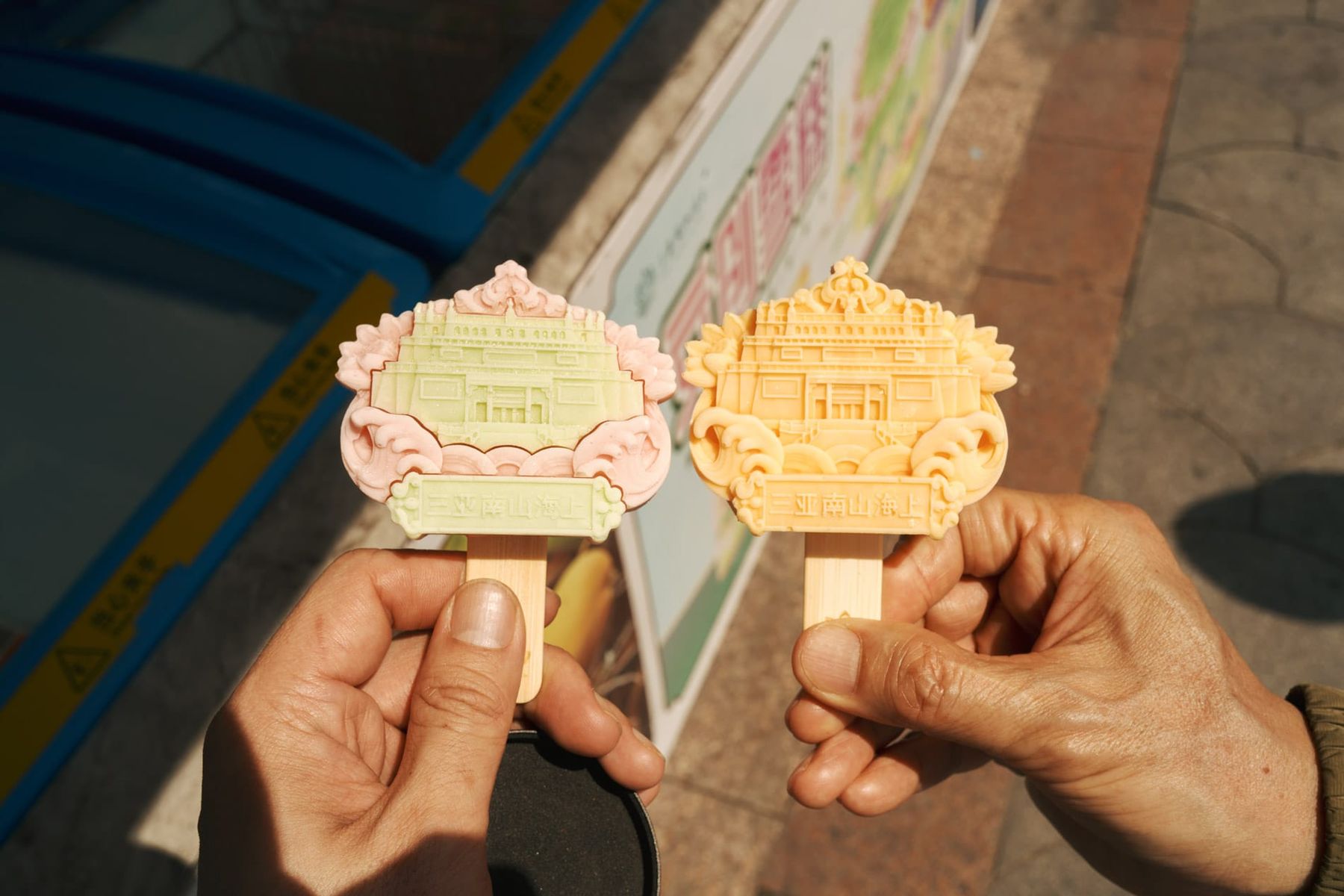 Two hands holding popsicles shaped like Buddhist temples, one pink and green, the other orange.